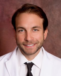 Dr Hromadka in lab coat and tie in front of red background